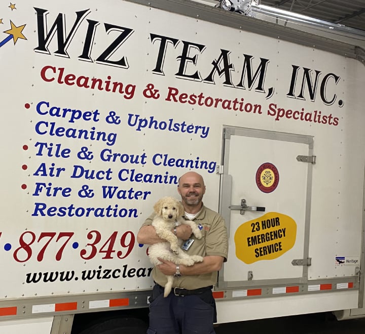 Owner of Wiz Team, Inc. holding a dog in front of a service vehicle displaying cleaning and restoration specialties, including carpet, upholstery, tile, and water restoration services.