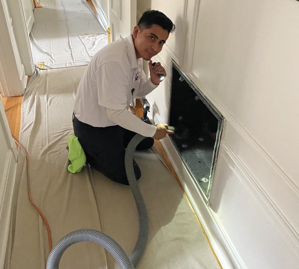 Technician performing air duct cleaning service, using a vacuum hose to remove dust and allergens, with protective covering on the floor in a residential setting.