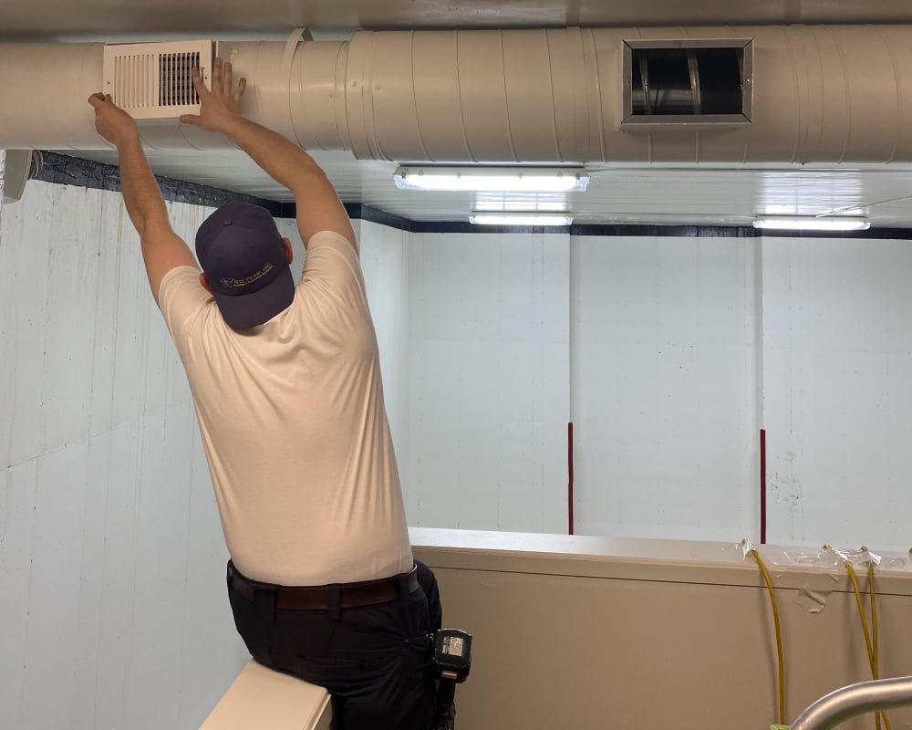 Technician performing air duct cleaning, reaching for a vent in a commercial setting, emphasizing HVAC maintenance and indoor air quality improvement.