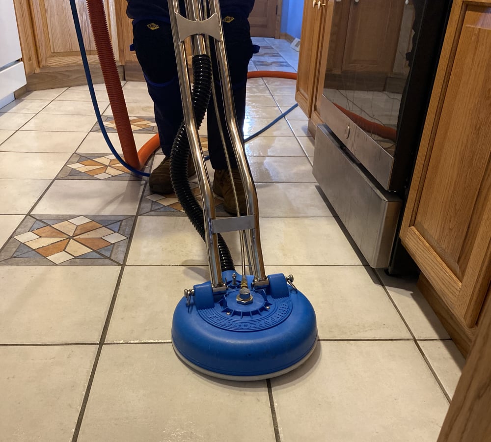 Professional tile and grout cleaning in a kitchen, featuring a worker using specialized equipment on patterned tile flooring, emphasizing cleanliness and maintenance.
