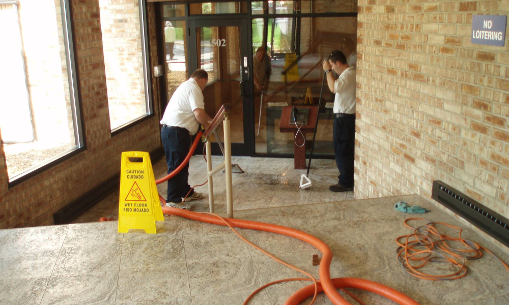Professional cleaners using equipment to maintain tile flooring in a commercial space, with a caution sign indicating a wet floor.