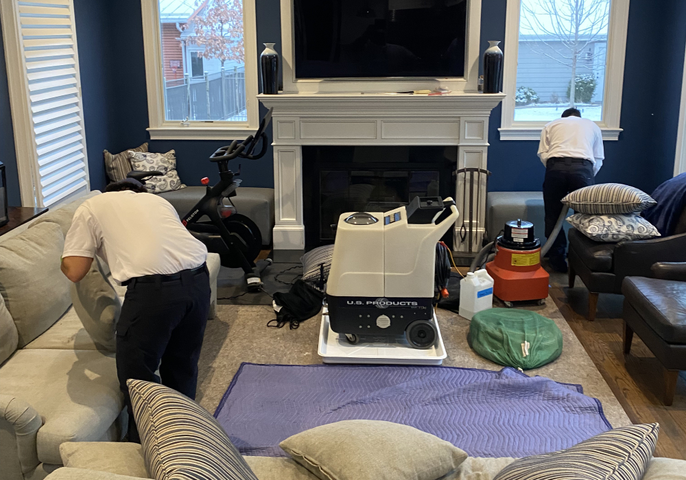 Two technicians cleaning upholstery in a living room, featuring a cleaning machine and vacuum, surrounded by furniture and decorative pillows, emphasizing professional upholstery care.