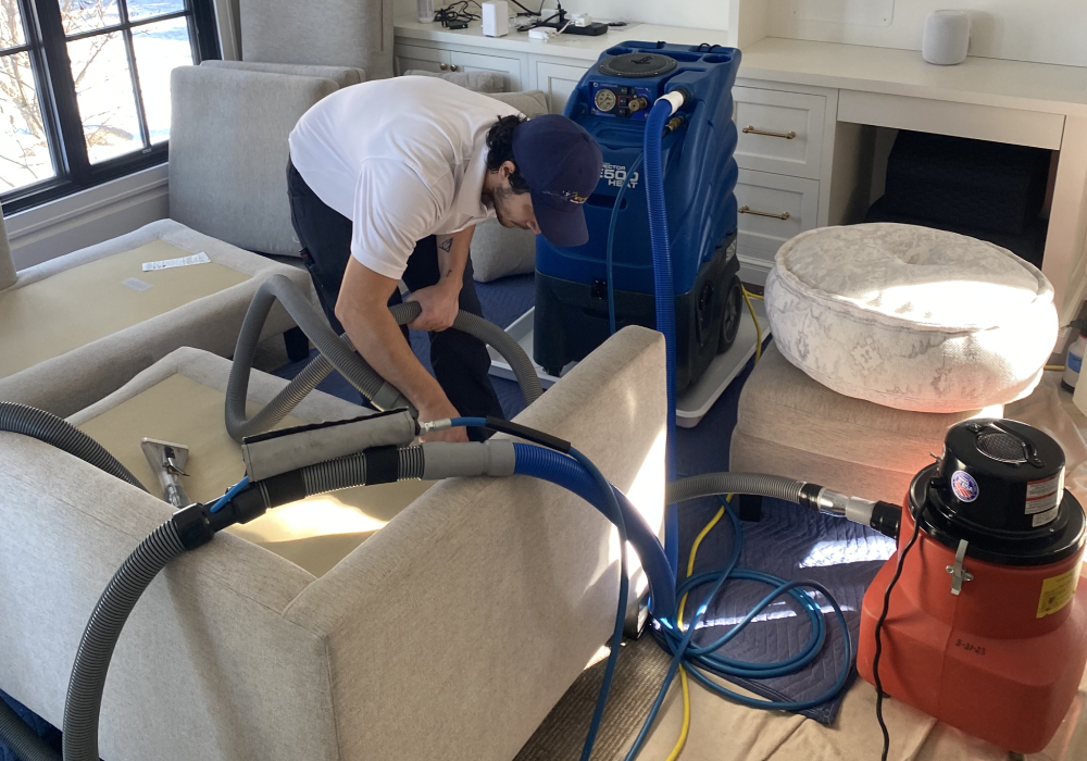 Upholstery cleaning in progress, technician using specialized equipment to clean fabric sofas, with cleaning machines and hoses visible in a well-lit living room setting.