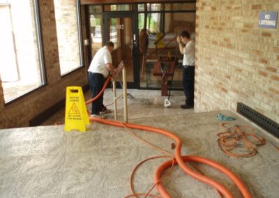 Two professionals performing tile and grout cleaning using steam equipment, with caution sign visible, in a well-lit interior space.