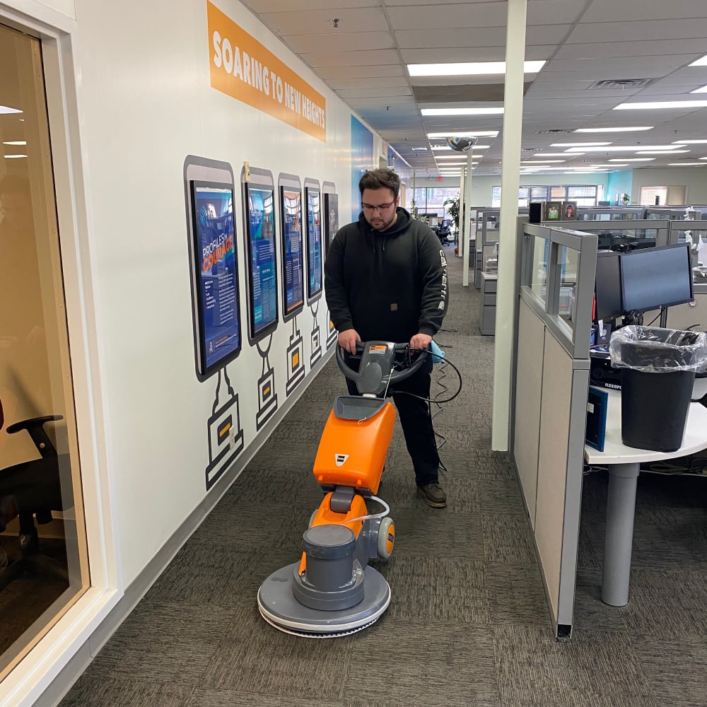 Person operating a floor cleaning machine in an office corridor, illustrating professional office cleaning services by Wiz Team Inc.