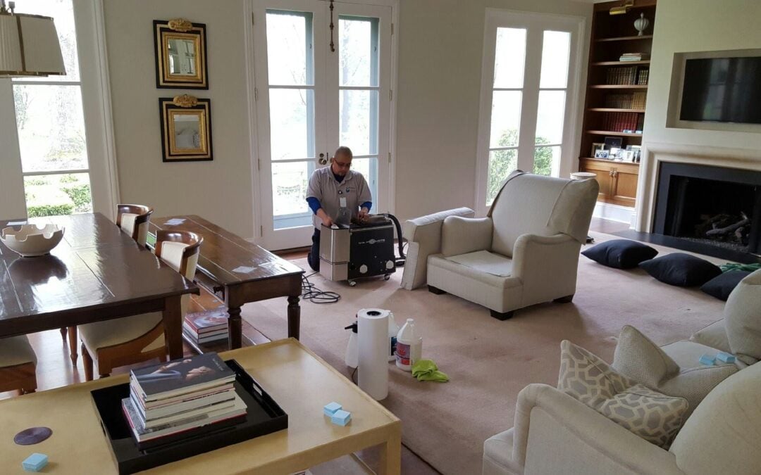 Man operating a carpet steam cleaning machine in a well-lit living room, surrounded by furniture and cleaning supplies, highlighting effective carpet maintenance methods.