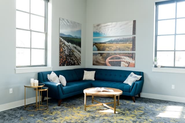 Cozy living room featuring a blue sectional sofa, decorative pillows, a round coffee table with a book, and large windows with scenic wall art, emphasizing cleanliness and comfort in home environments.