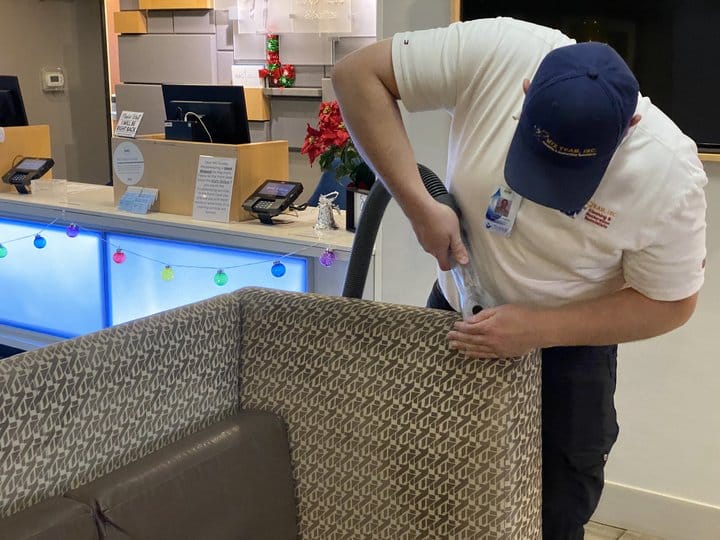 Professional cleaner using upholstery cleaning equipment on patterned couch in busy reception area, emphasizing effective cleaning solutions for households.