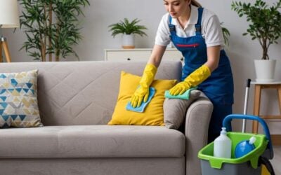 Woman in blue apron and yellow gloves cleaning a light-colored sofa with eco-friendly cleaning supplies, emphasizing professional upholstery care for improved indoor air quality.